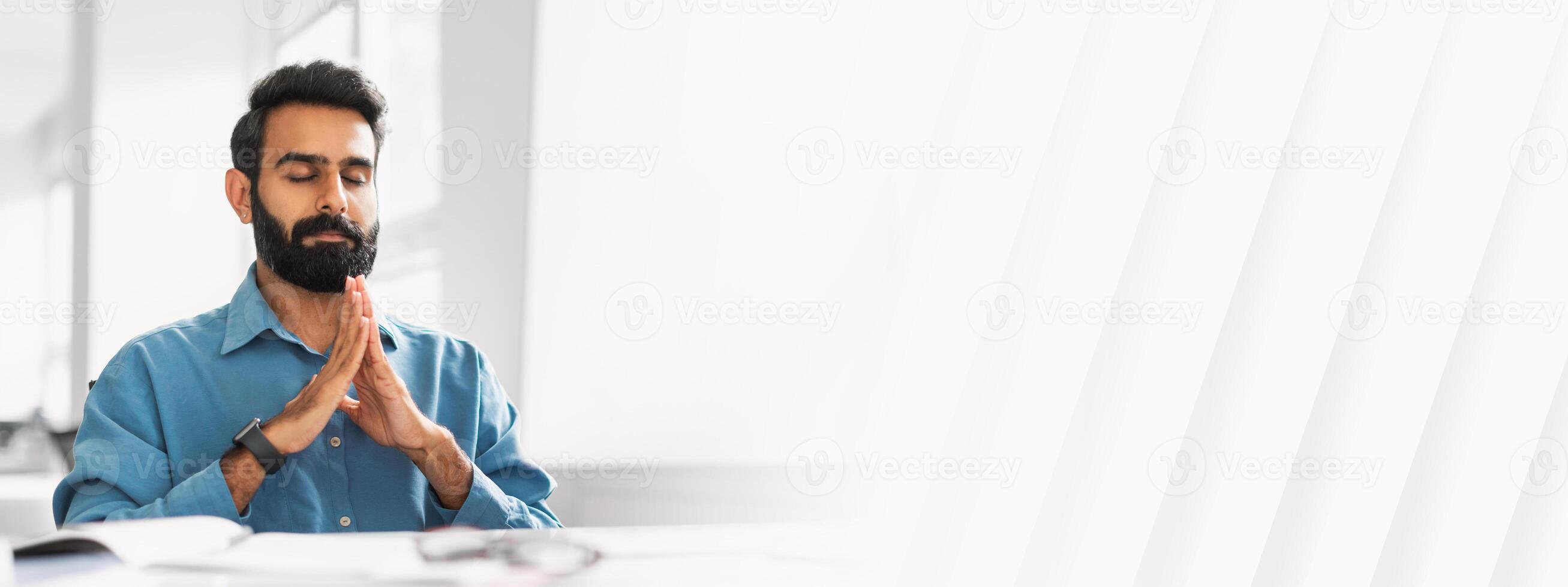 Indian businessman with closed eyes seeking tranquility through meditation, hands pressed together, at his bright office workspace, free space photo
