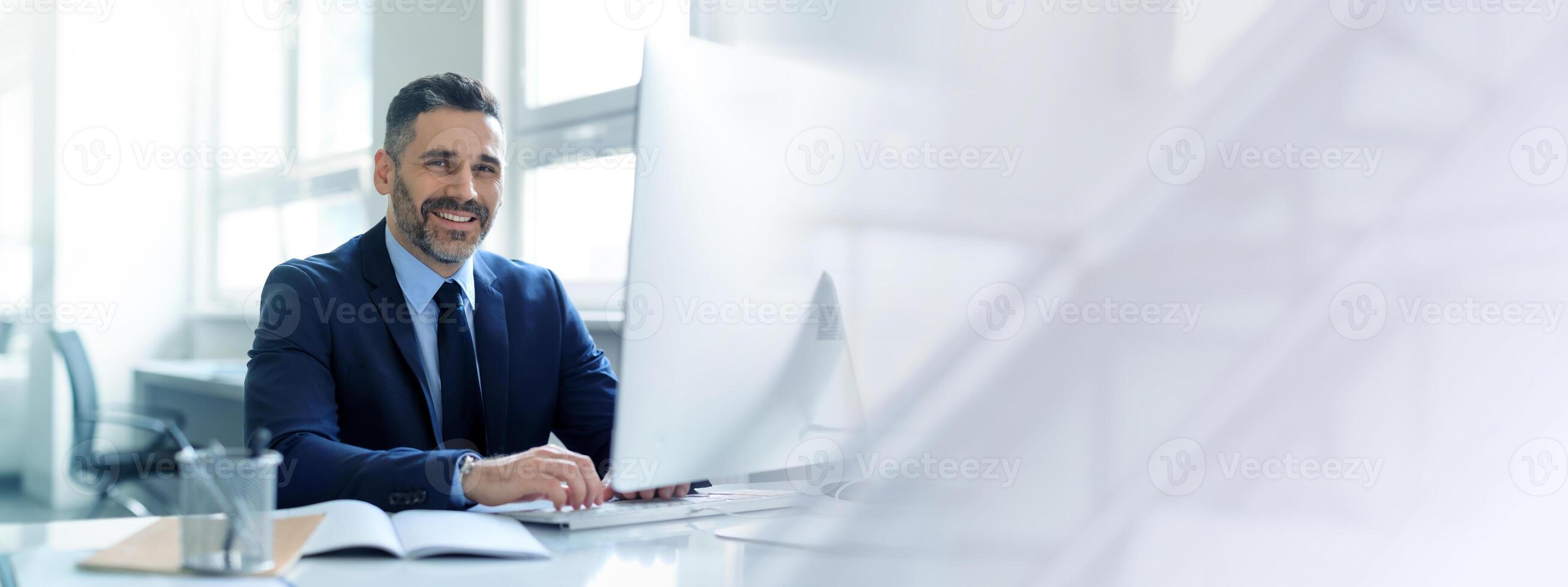 A middle aged businessman with a mature look is focused on his work at a sleek office desk. He exudes confidence while engaging with his computer. Bright natural light illuminates the space. photo