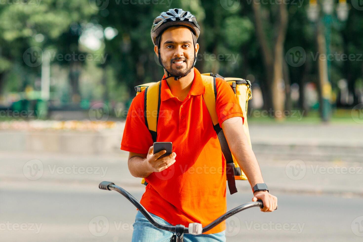 Cyclist with delivery. Happy courier with yellow backpack, smart watch and smartphone, sits on bicycle in city, copy space photo