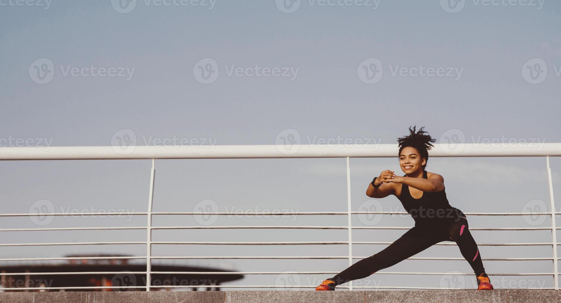 Exercises for health and slim body. Smiling african american girl in sport uniform squats and stretches at stadium on sky background, free space photo