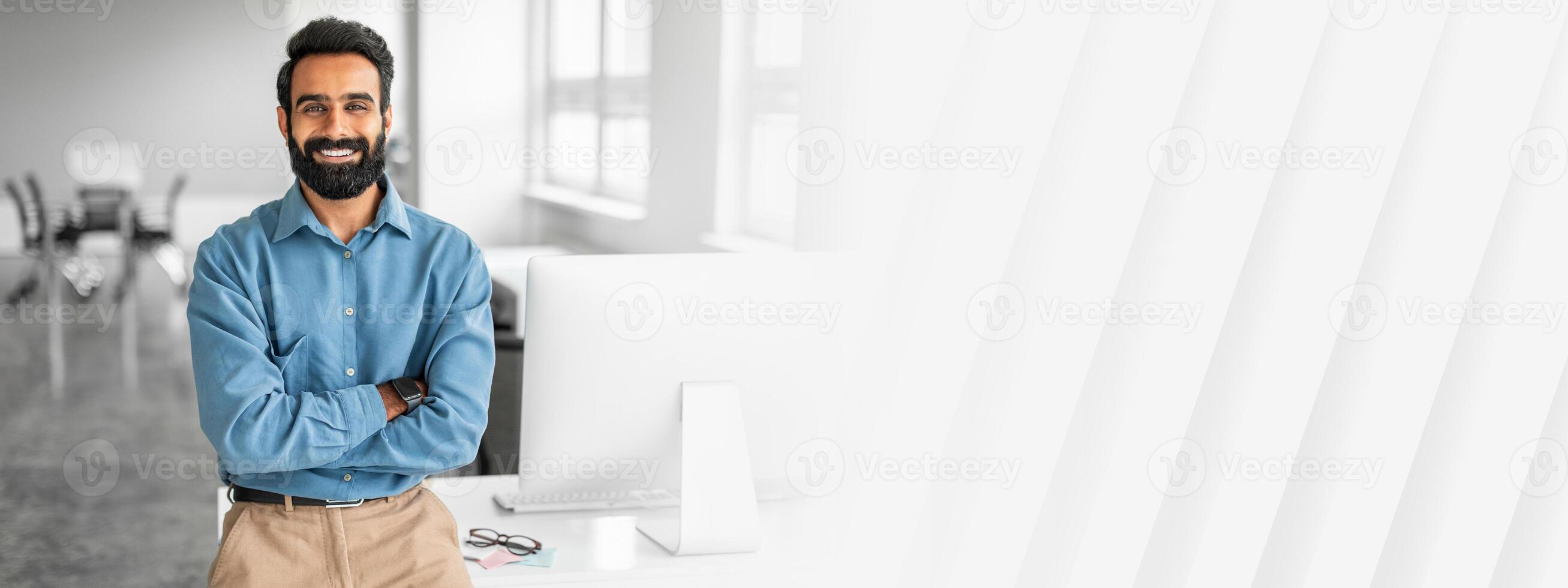 Happy indian businessman standing by his workplace with folded arms, smiling at camera, planning investment, thinking of new startup or project, enjoying business success photo