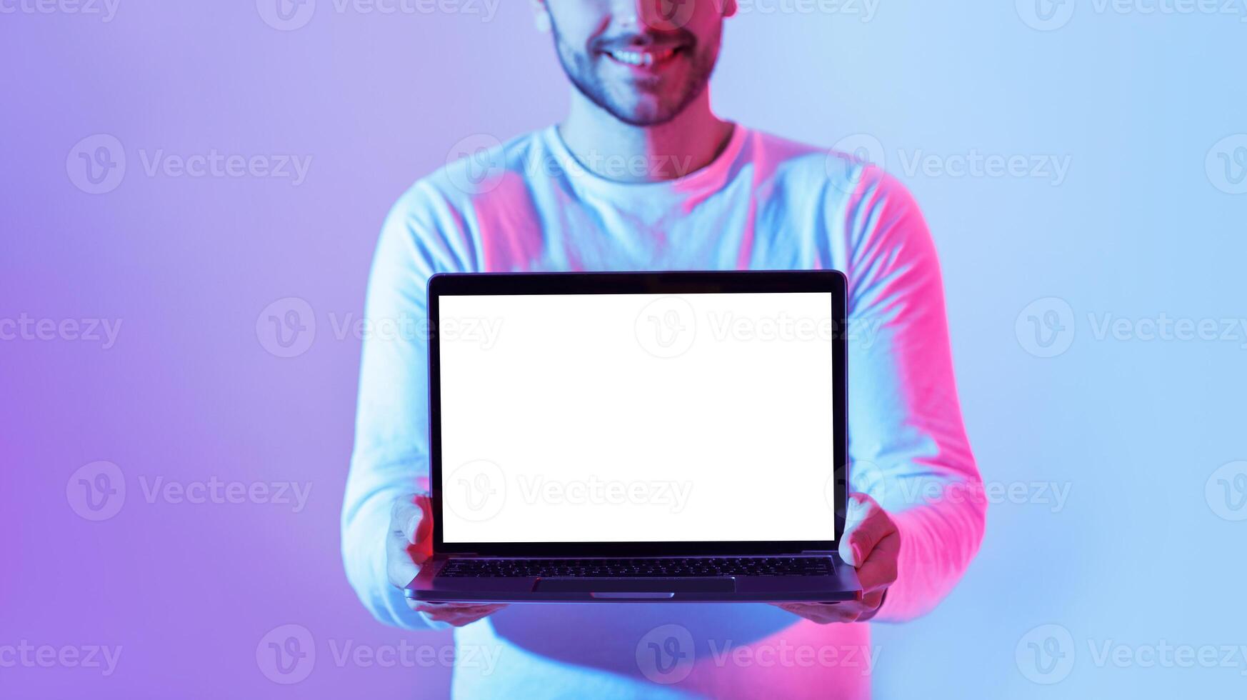 A young man holds a blank laptop in front of him, smiling brightly. The background features vibrant blue and purple lighting, creating a modern and energetic atmosphere. photo