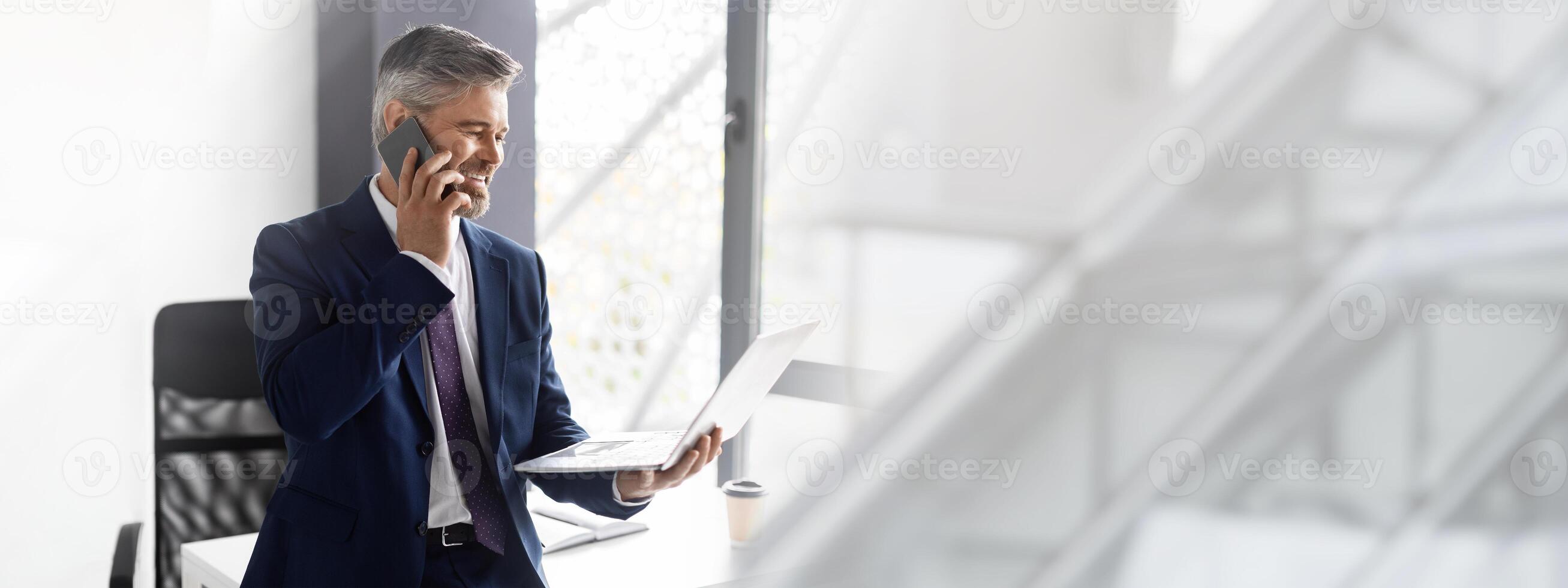 Middle Aged Businessman In Suit Talking On Cellphone And Using Laptop At Office, Mature Boss Looking At Computer Screen And Having Business Call With Client While Leaning At Desk, Copy Space photo
