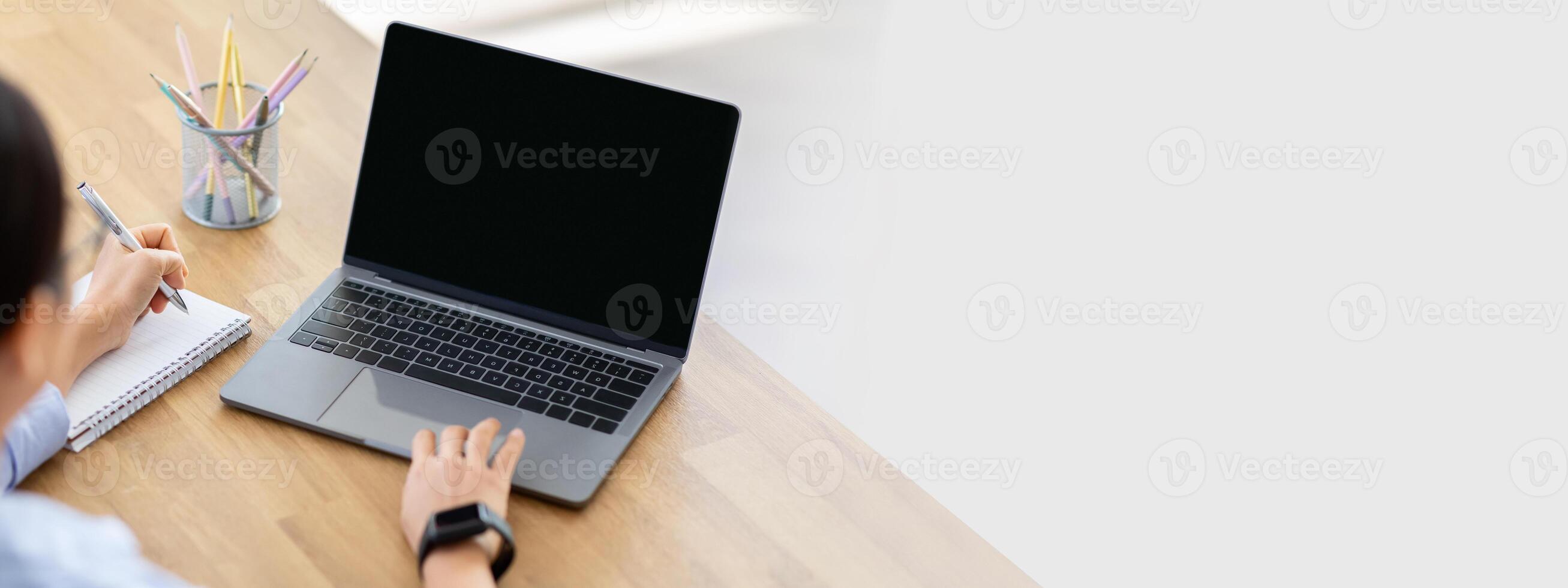 High angle view asian woman using laptop computer with blank screen mockup copy space, taking notes in notepad, attending webinar, have online lesson, home interior. E-education photo