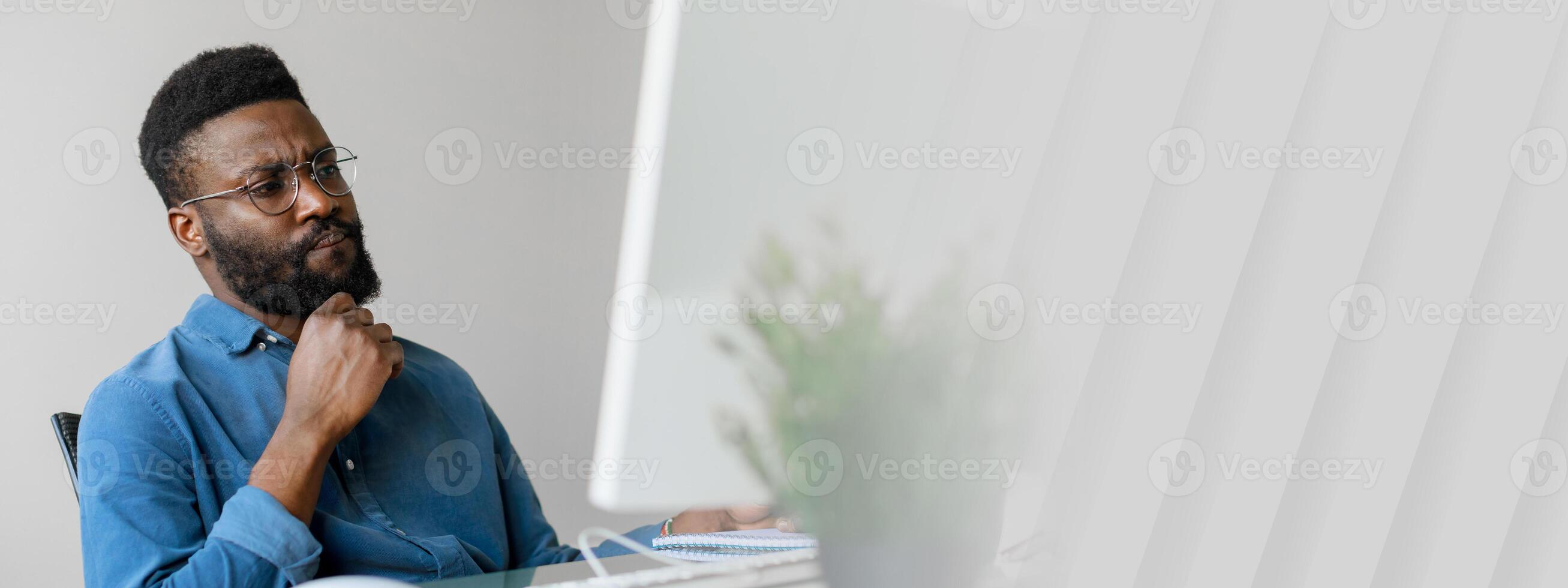 A young African American entrepreneur sits thoughtfully at a desk in a contemporary office. He is engaged in work on a computer, exemplifying the modern professional lifestyle, copy space photo