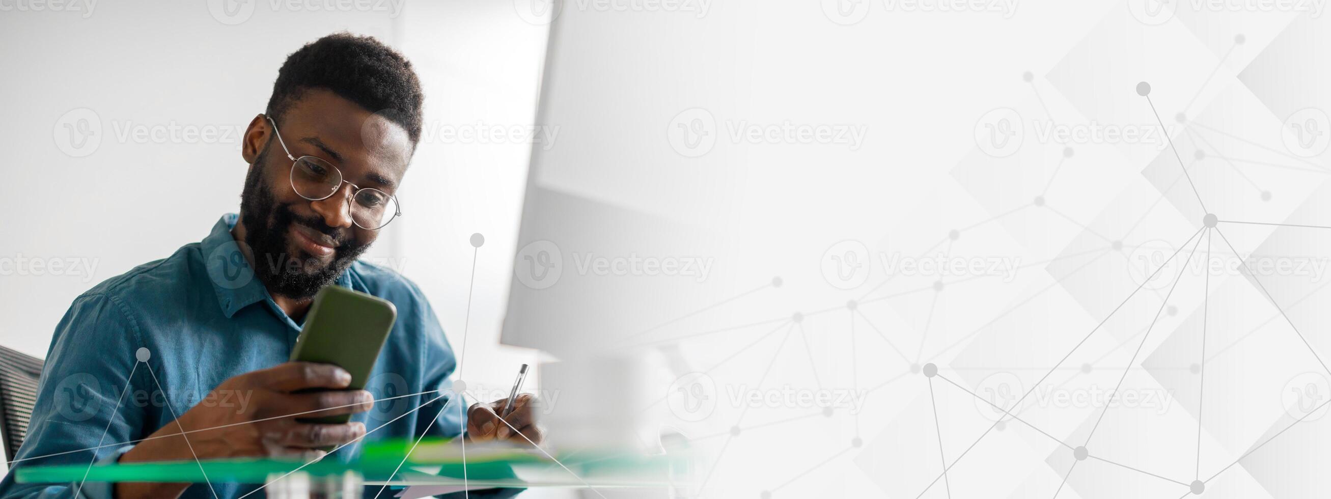 A young African American entrepreneur actively works in his office, using a smartphone to manage tasks and jotting down notes. The modern workspace showcases a lifestyle of success and ambition. photo
