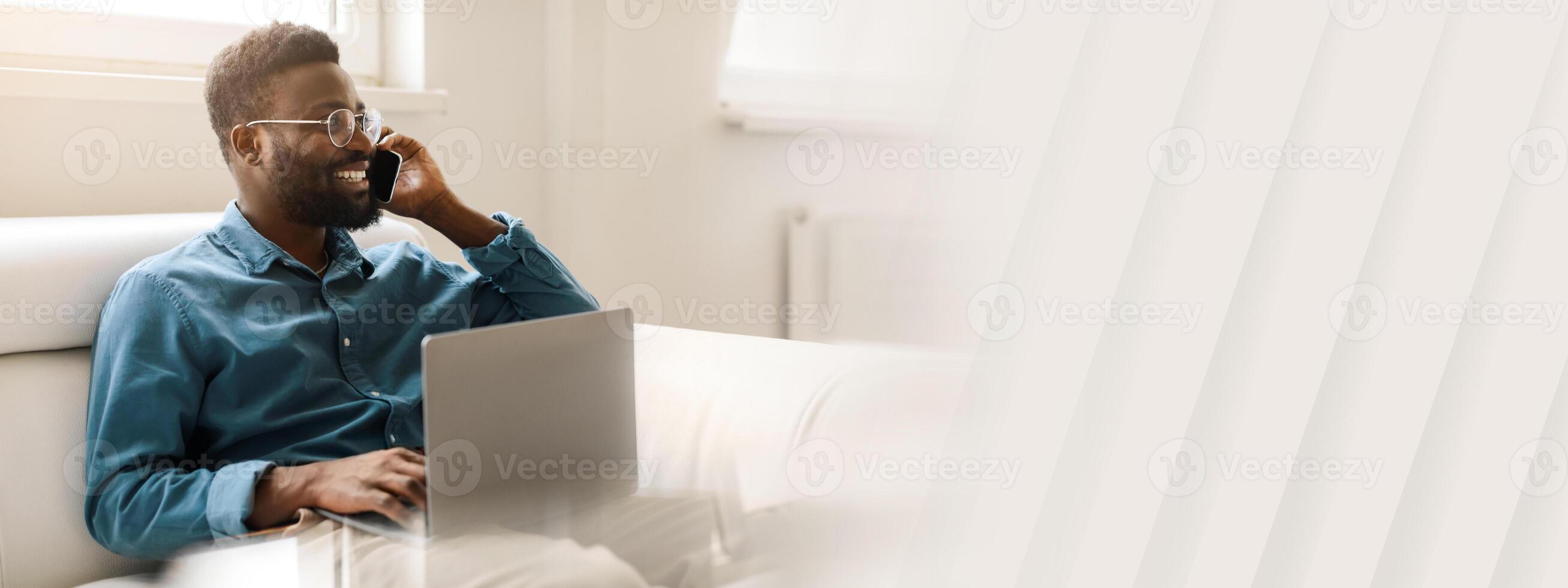A young African American man sits on a sofa in an office, engaged in a phone conversation while working on his laptop. He embodies success and a modern entrepreneurial lifestyle, copy space photo