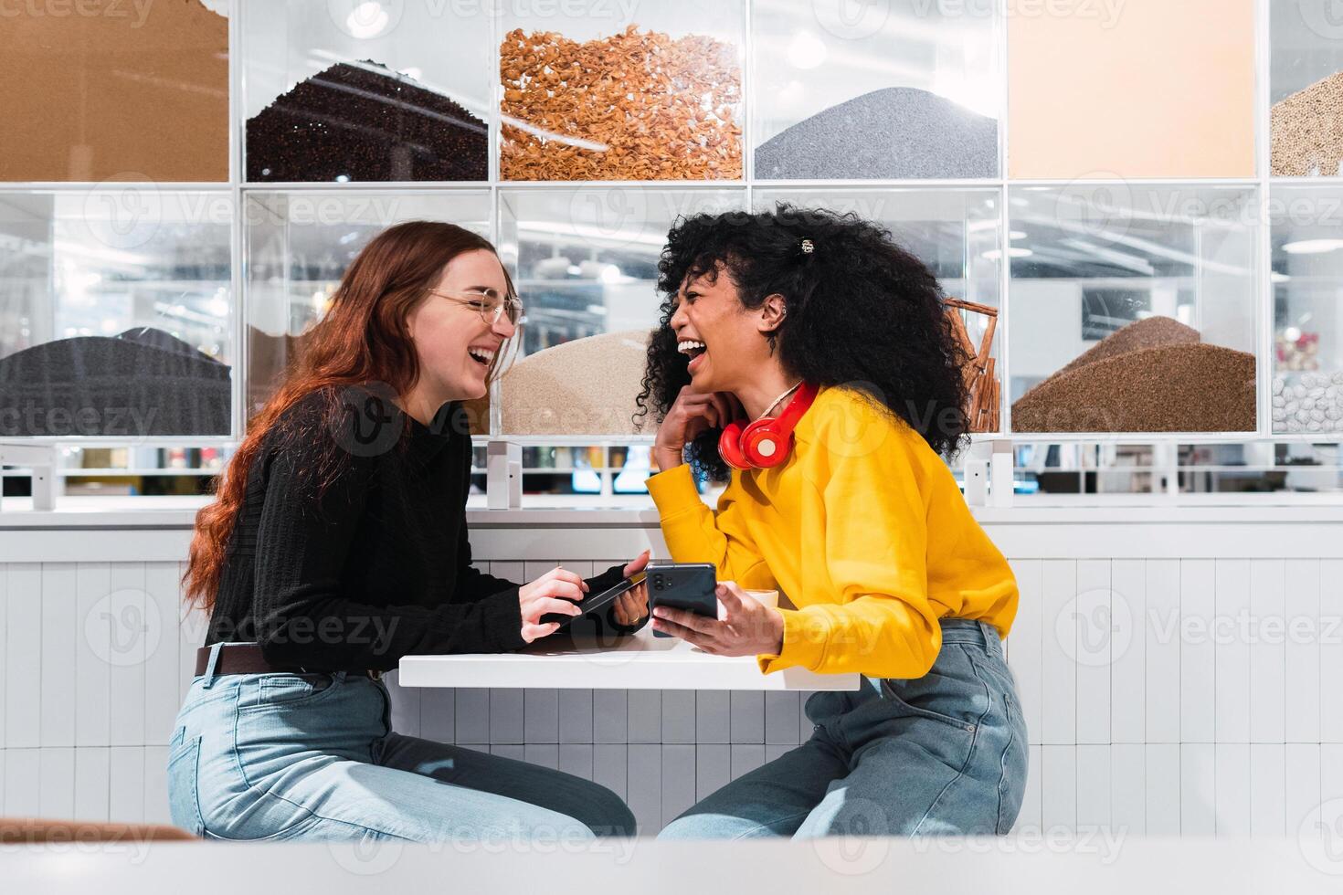 Positive multiethnic female friends using smartphones and laughing in cafe photo