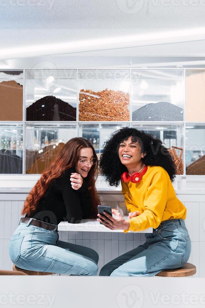 Positive multiethnic female friends using smartphones and laughing in cafe photo
