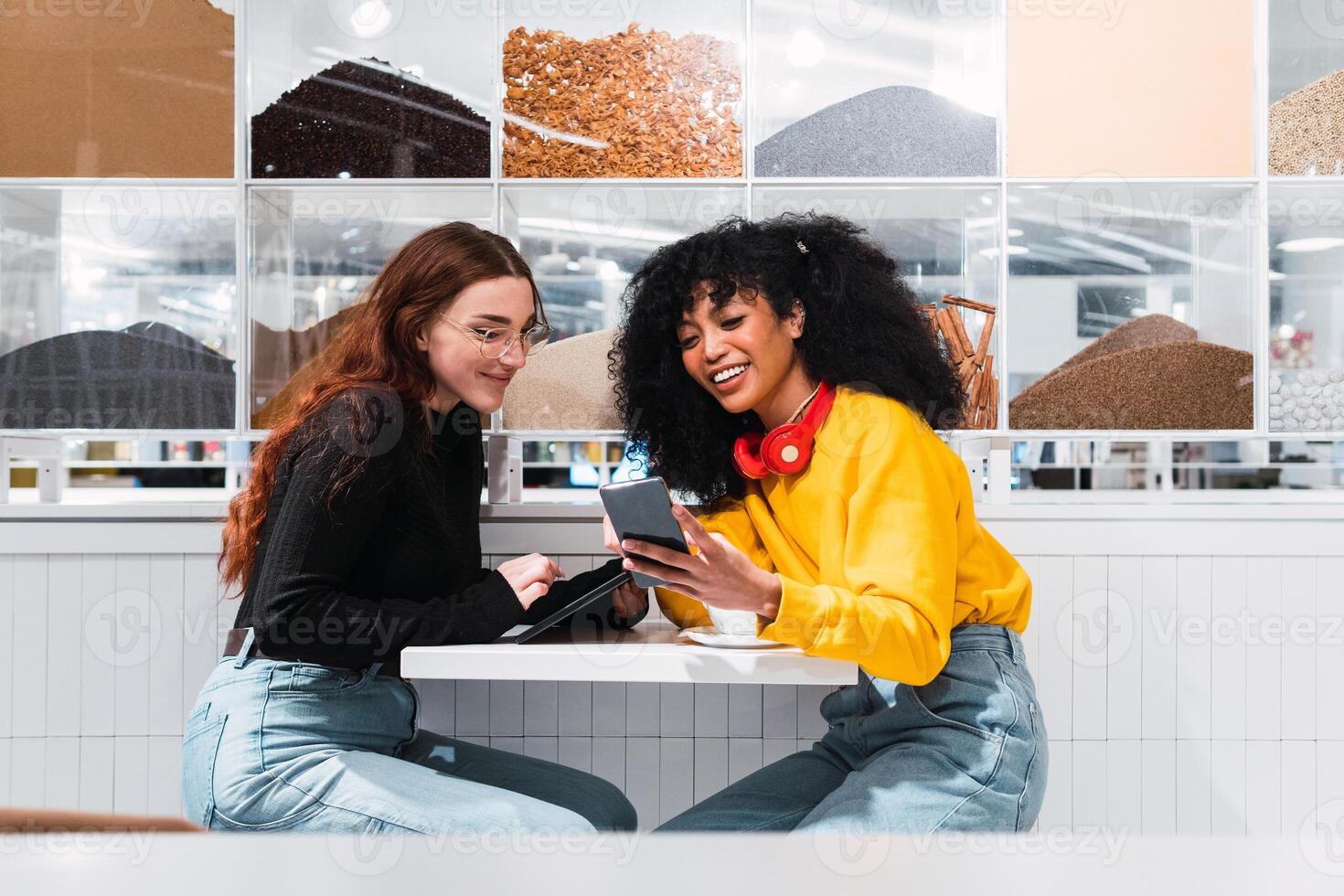 Positive multiethnic female friends using smartphones and laughing in cafe photo