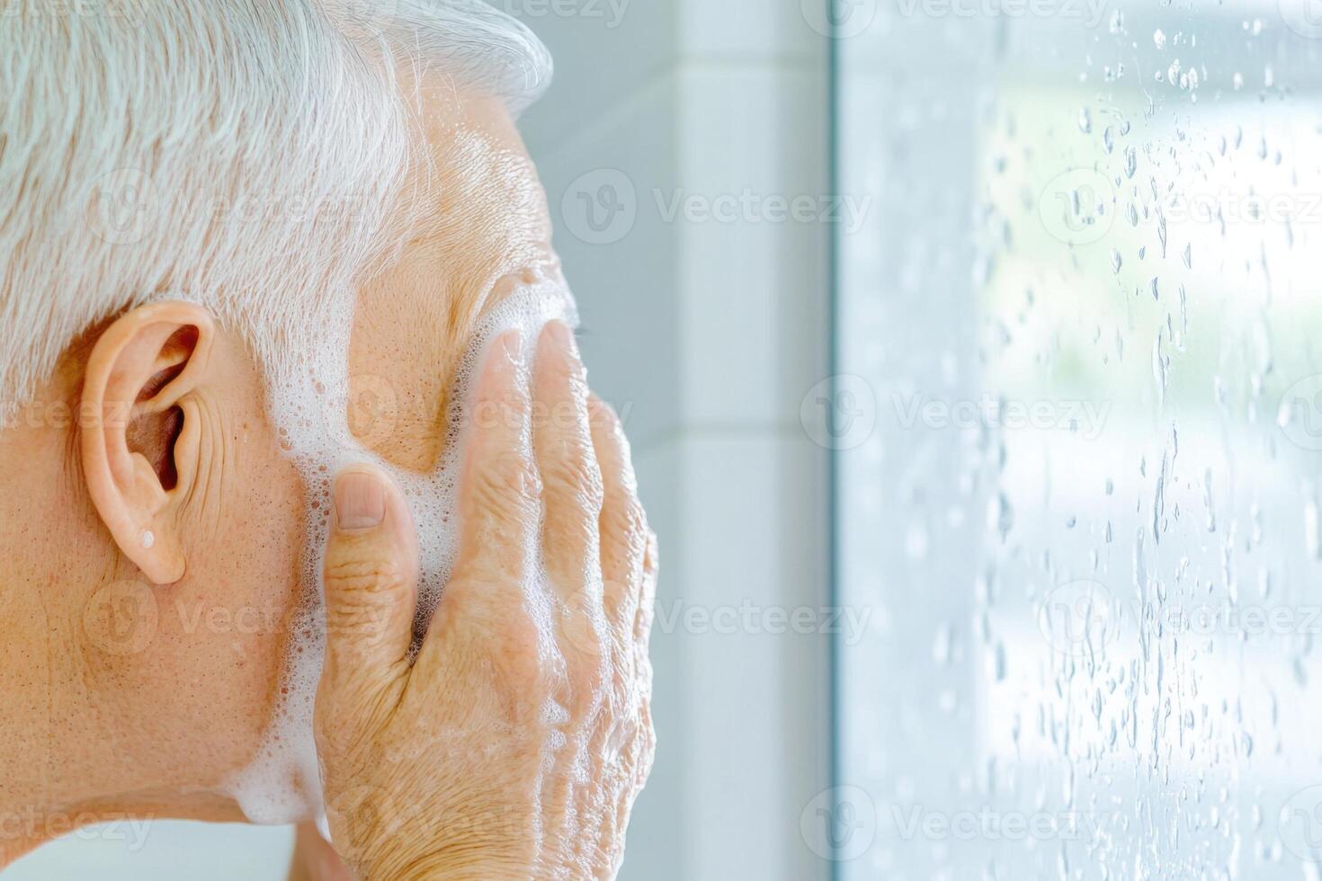 Elderly Asian man washing his face with cold water in a modern bathroom with natural light streaming through a frosted window photo