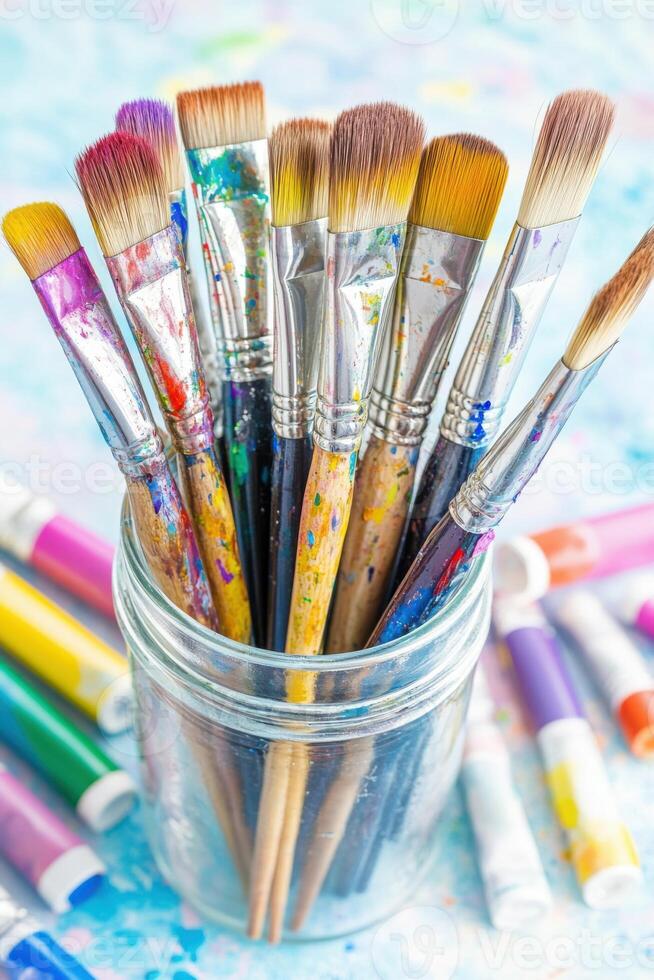Well-used artist paintbrushes with dried paint in a glass jar and paint tubes displayed on a colorful surface, creating an artistic and creative environment photo