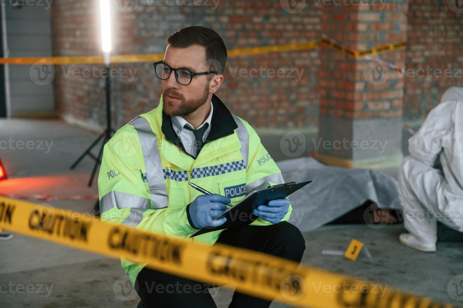 Man in glasses is holding notepad and pen. Detectives are collecting evidence in a crime scene near dead body photo