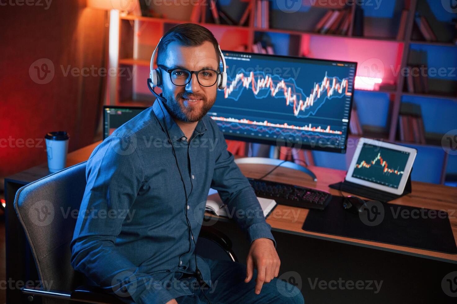 Sitting and looking ,a,t the camera. Stock trader is working by pc indoors. Conception of exchange and money photo