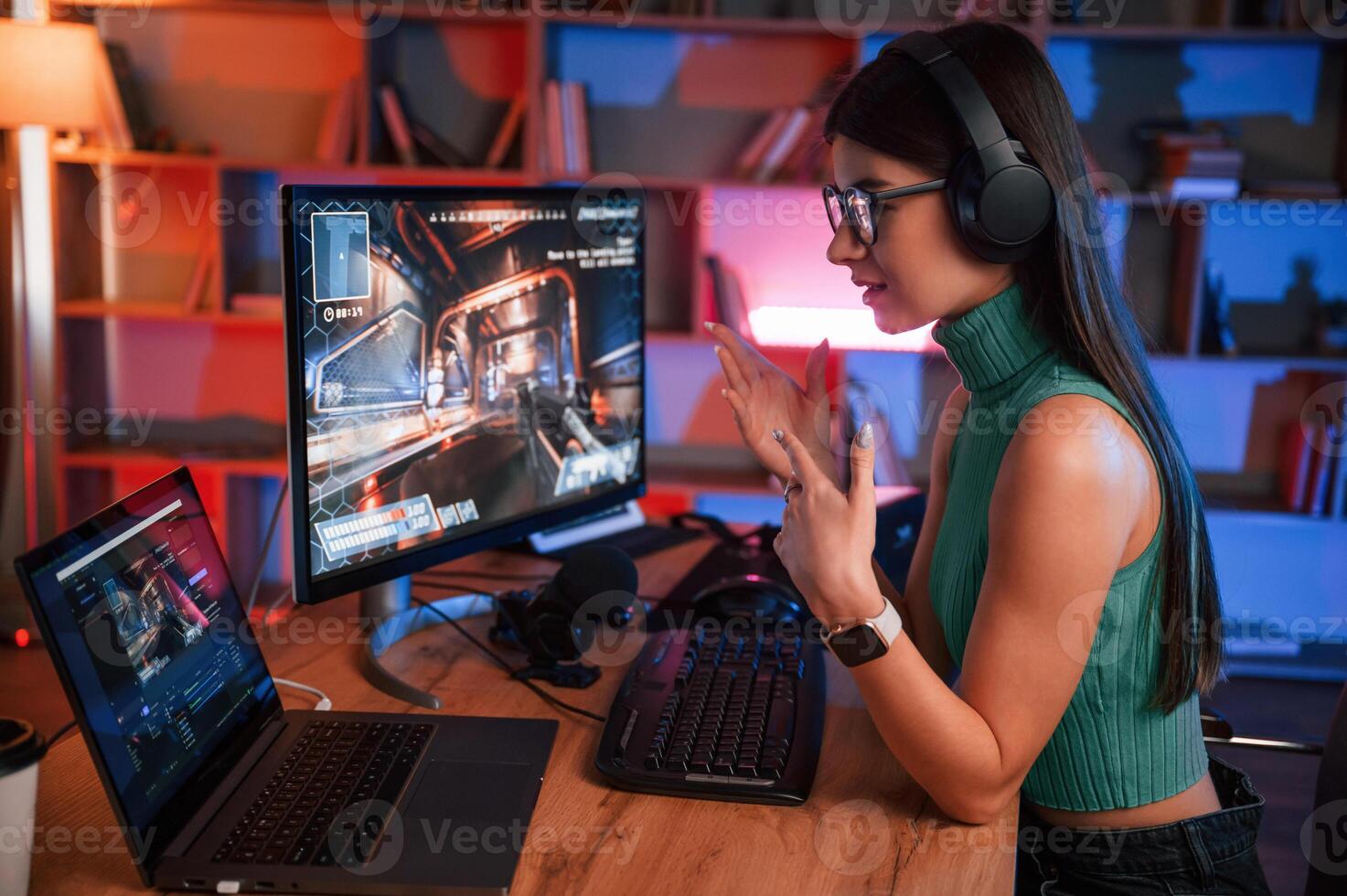 In headphones. Young pretty female, gamer is playing shooter. Neon lighting photo