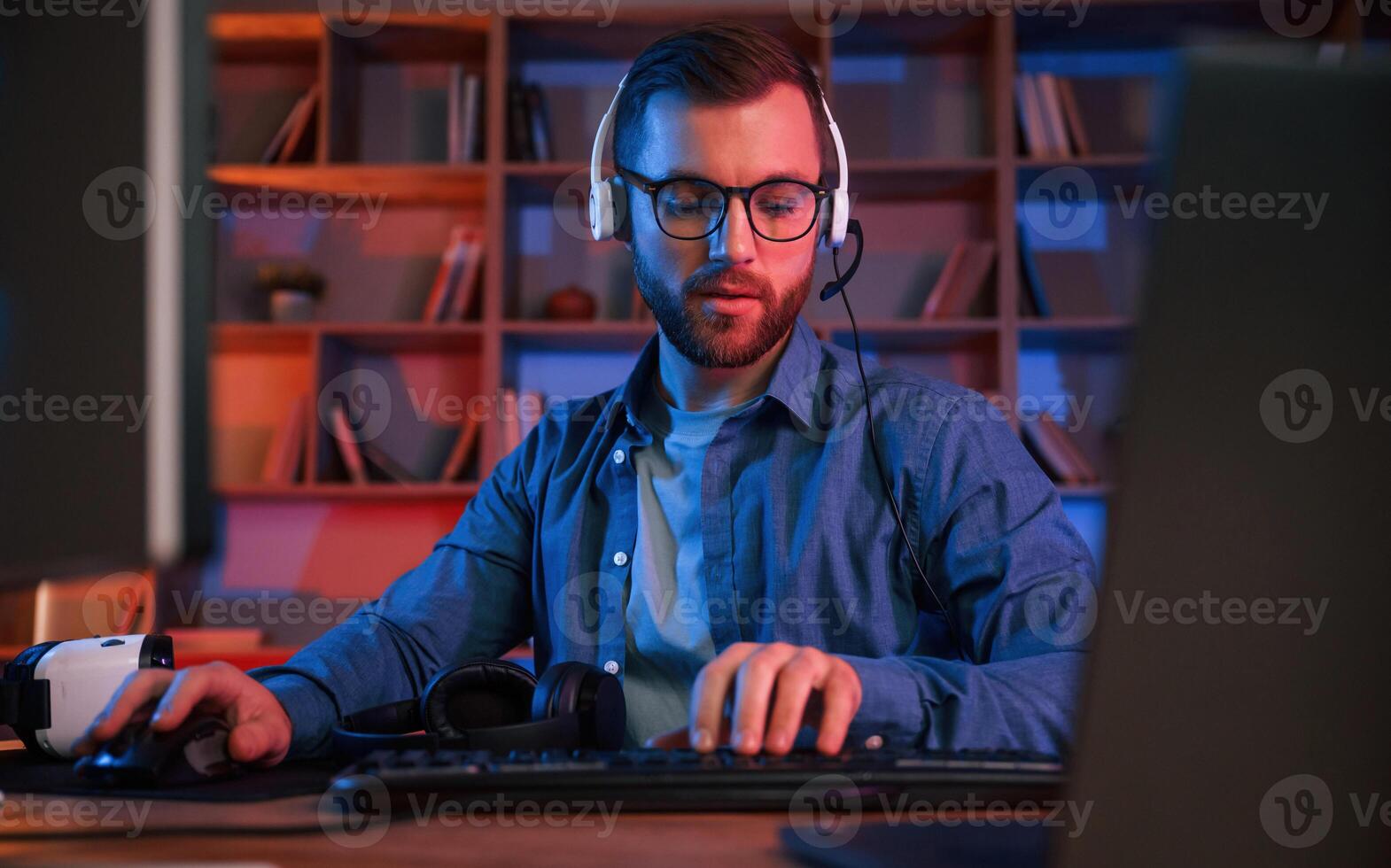Front, view. Man is sitting by a computer indoors at home. Neon lighting photo