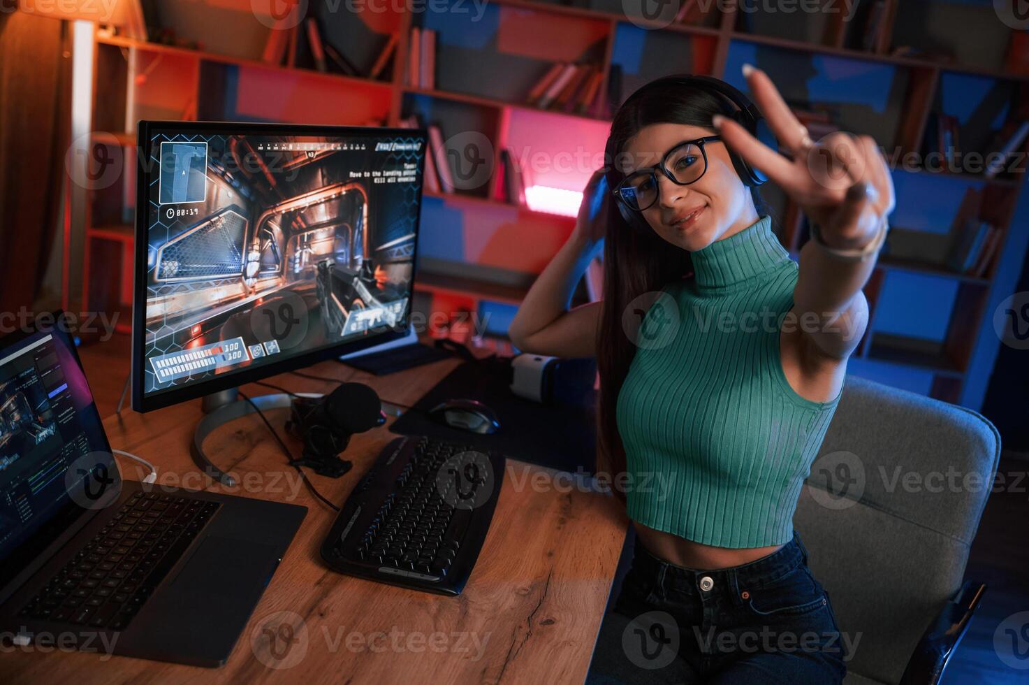 Smiling, showing,, victory gesture. Young pretty female gamer is playing shooter. Neon lighting photo