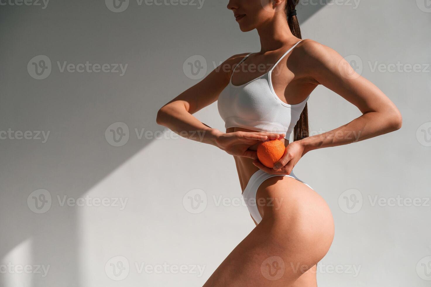 Orange fruit is in the hands.. Young caucasian woman with slim body shape is indoors in the studio photo