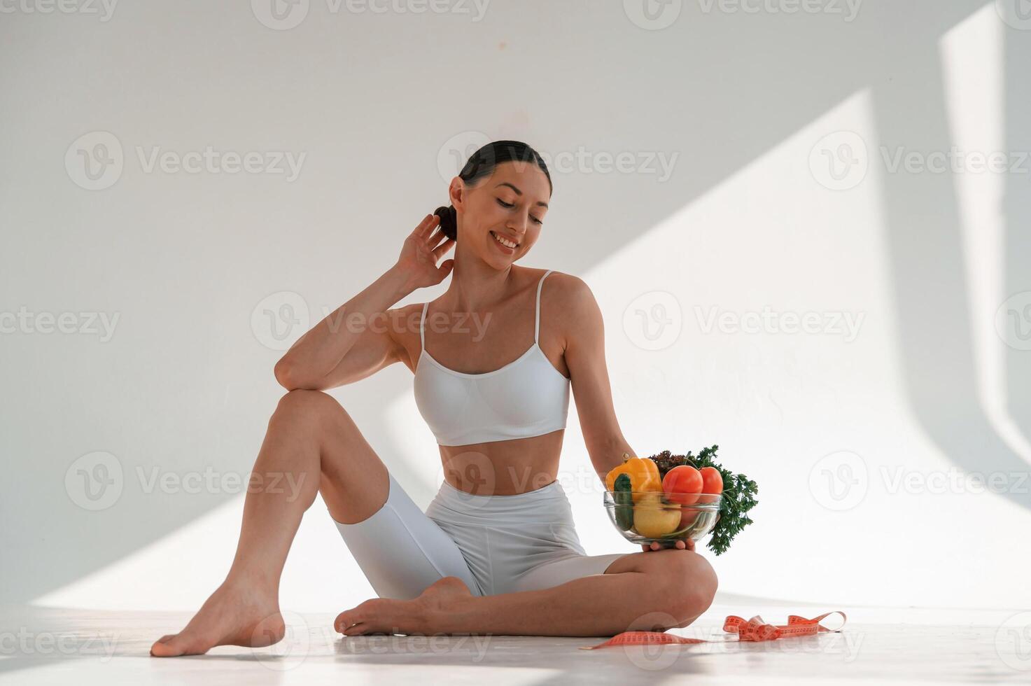 Sitting on the floor with healthy food. Young caucasian woman with slim body shape is indoors in the studio photo