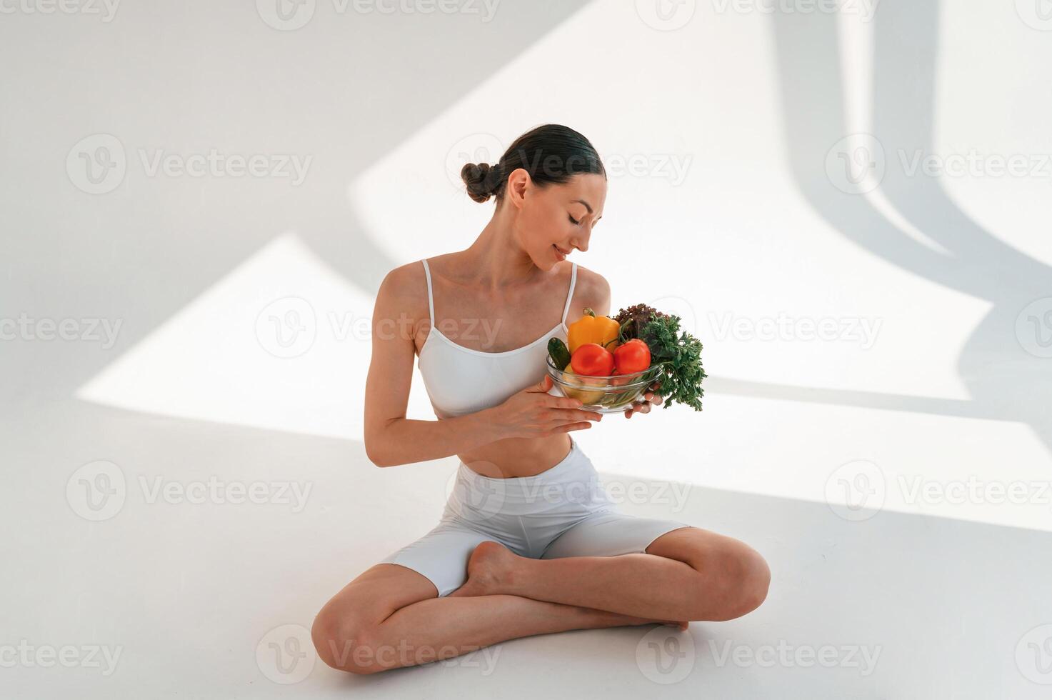 Looking at healthy food. Sitting on the floor. Young caucasian woman with slim body shape is indoors in the studio photo