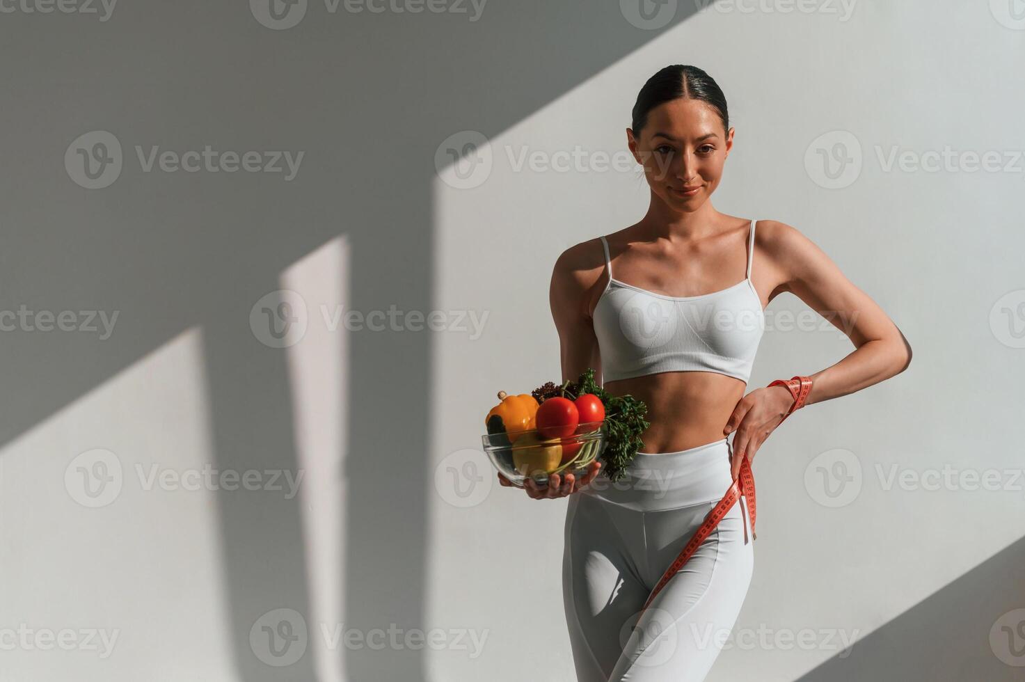 Holding a bowl of vegetables and fruits. Young caucasian woman with slim body shape is indoors in the studio photo