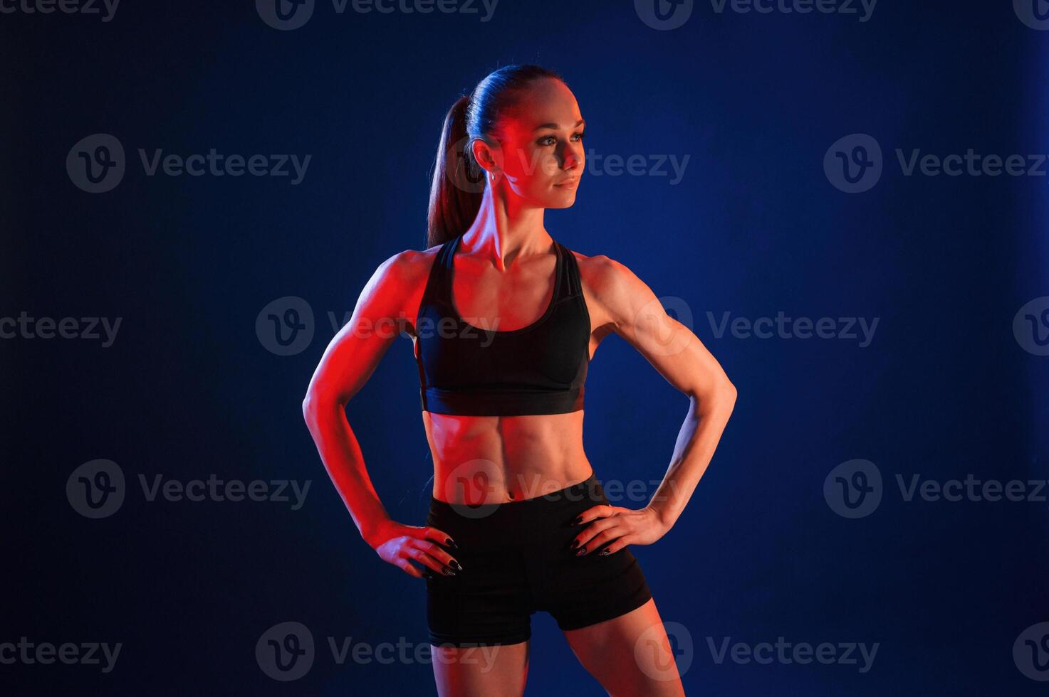 Standing, with the hands ,on waist. Beautiful muscular woman is indoors in the studio with neon lighting photo