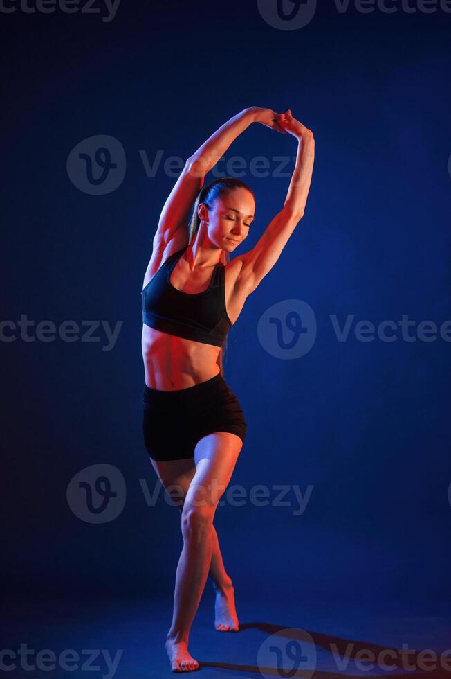 Sportive exercises, standing and stretching the hands. Beautiful muscular woman is indoors in the studio with neon lighting photo