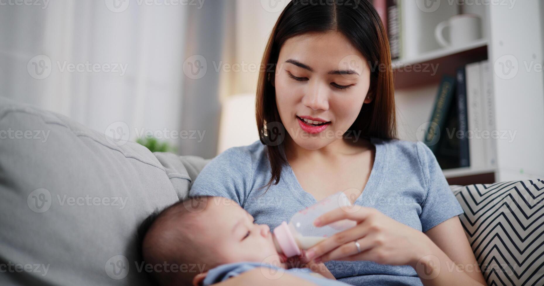 Beautiful mother gently holds her little son in her arms and feeds him with a bottle while sitting on a sofa in the living room, with an atmosphere full of love photo