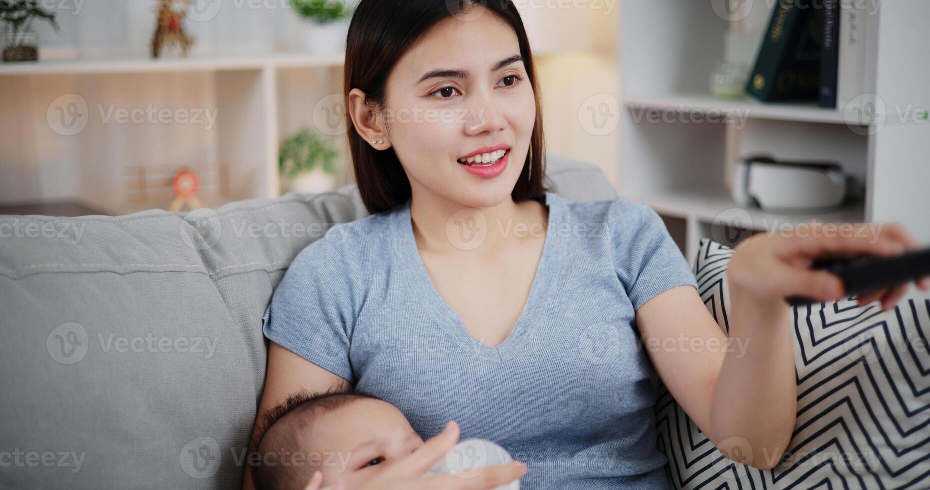 Portrait of A beautiful mother gently holds her little son in her arms, feeds him milk with a bottle, and uses the remote control to select channels.. photo