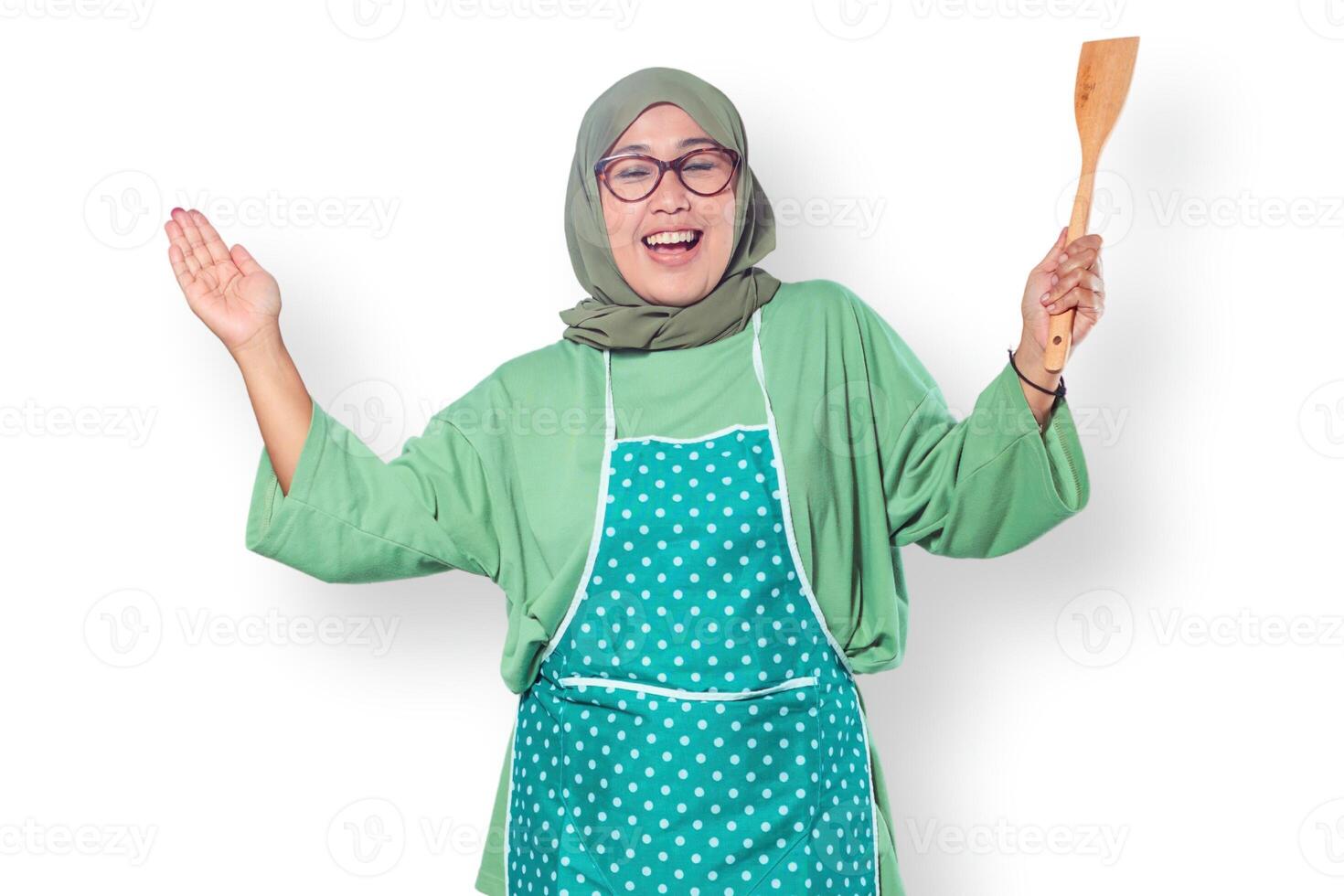 Cheerful Muslim woman chef wearing an apron, holding a spatula, and smiling confidently in a home kitchen setting photo