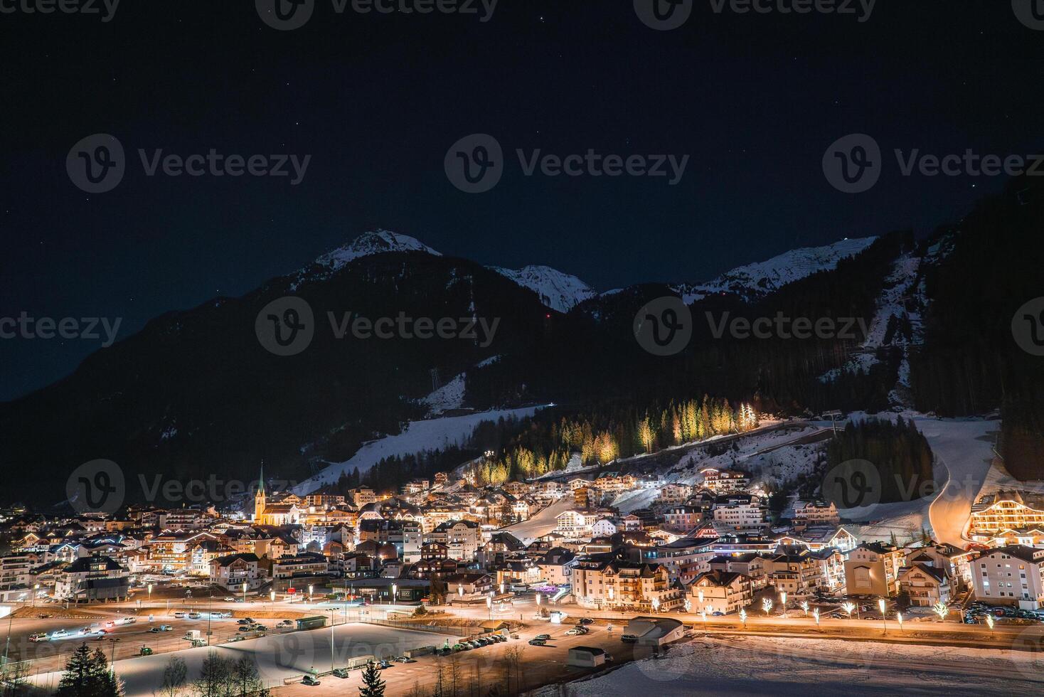 A well lit alpine village in Ischgl, Austria, at night, surrounded by snow covered mountains. A church steeple and illuminated slopes are visible. photo