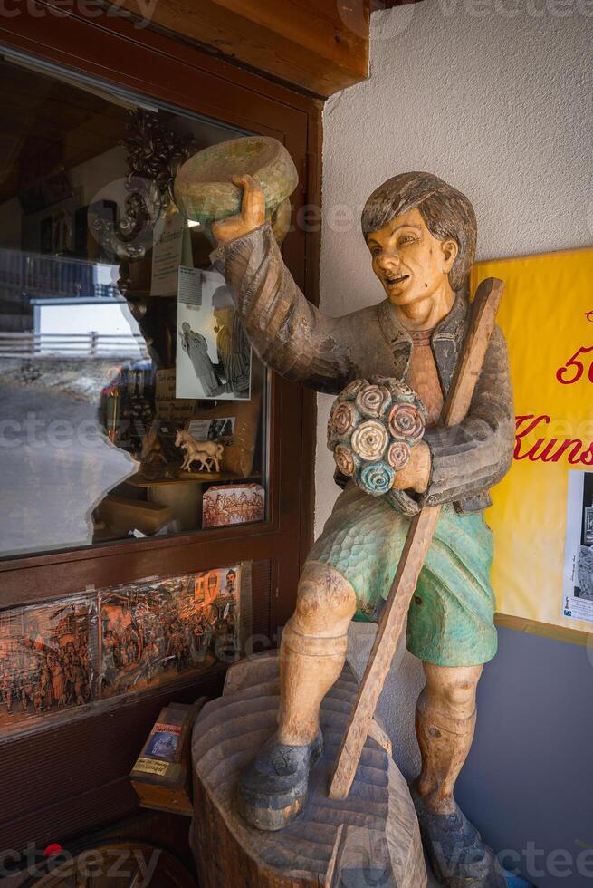 A wooden figure with green shorts holds a bowl and flowers near a shop window displaying figurines and artwork in Ischgl, Austria. photo