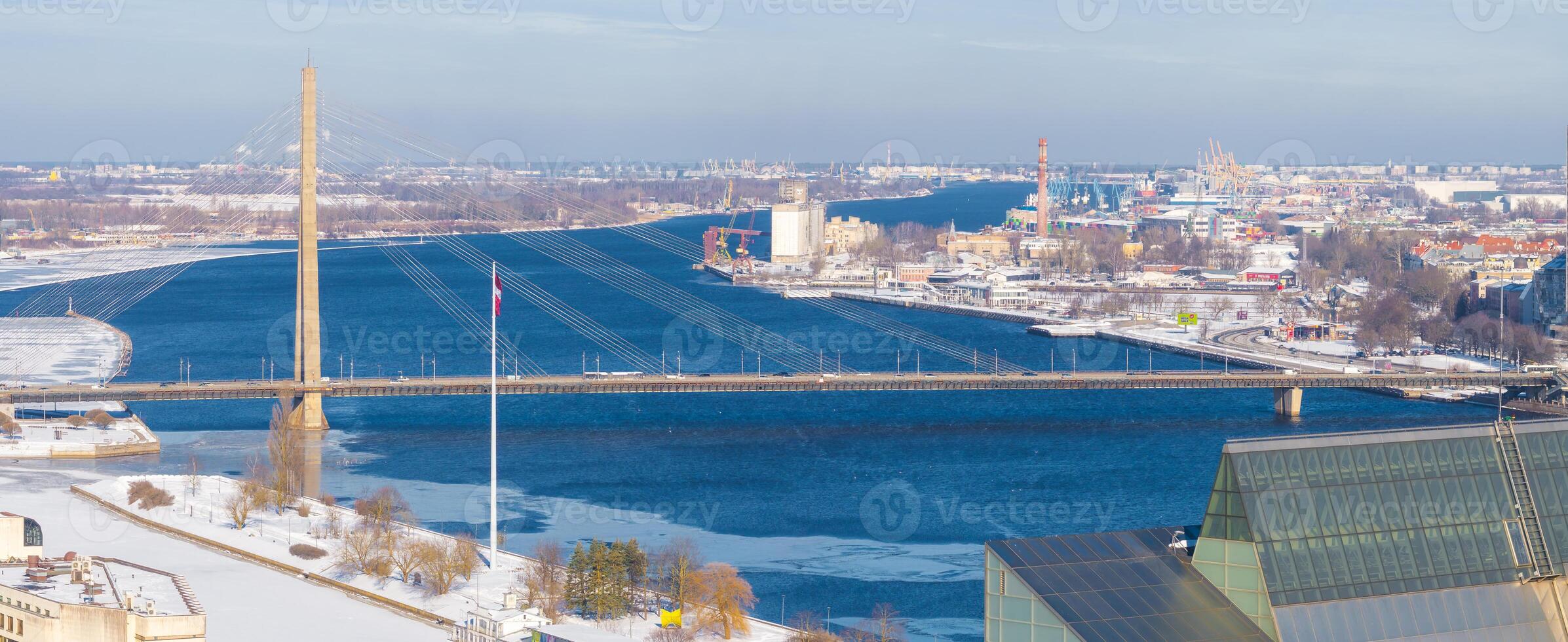Aerial view of Riga, Latvia, in winter, showing the Vansu Bridge over the Daugava River, the National Library, a frozen river, and industrial structures. photo