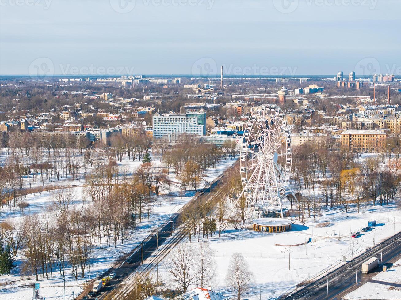 Aerial view of Riga, Latvia, featuring a Ferris wheel in a snowy park, urban buildings, tram tracks, and a clear sky with distant cityscape elements. photo