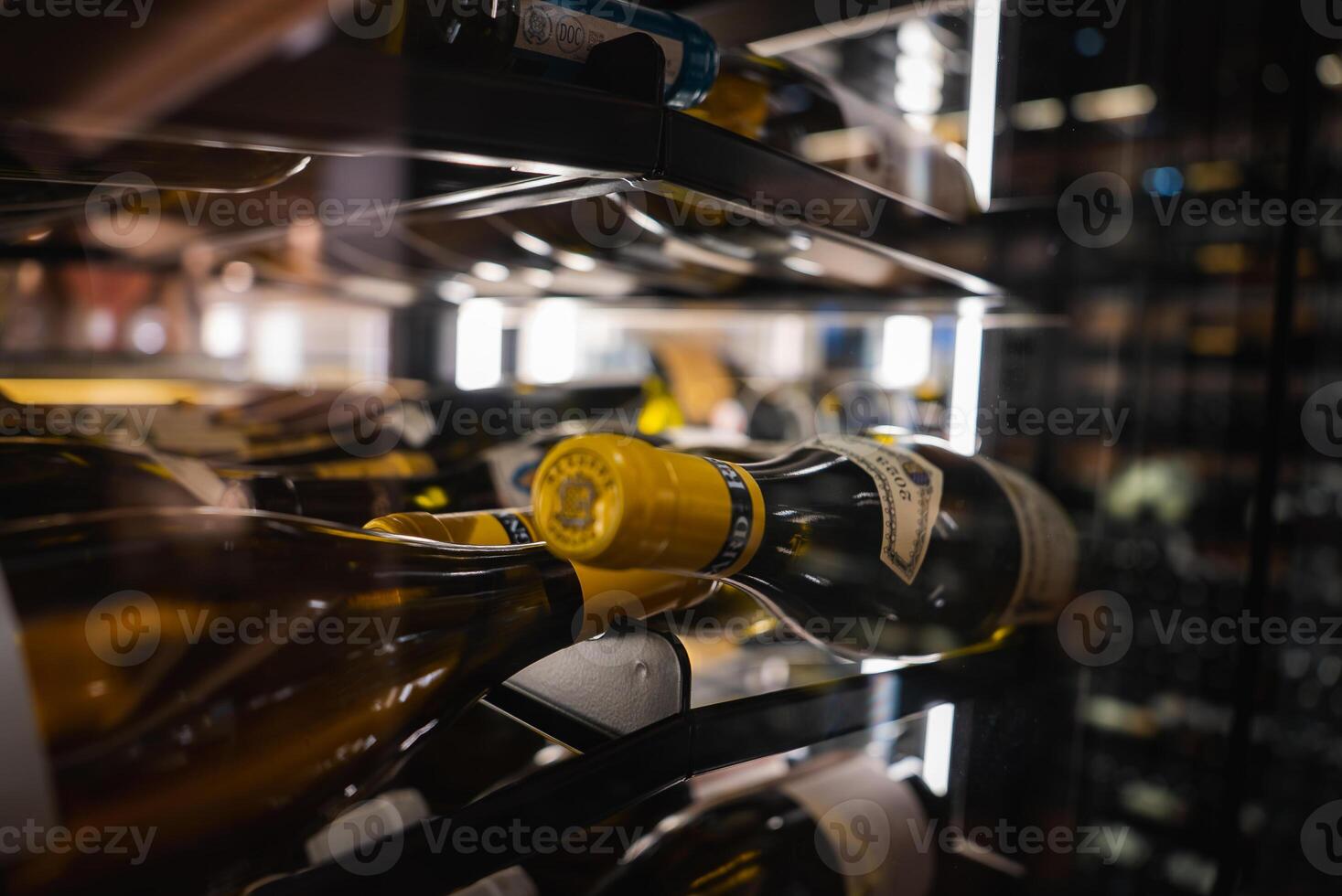 Horizontal wine bottles with yellow caps and detailed labels in a well lit, modern wine cellar with reflective surfaces and blurred racks. photo