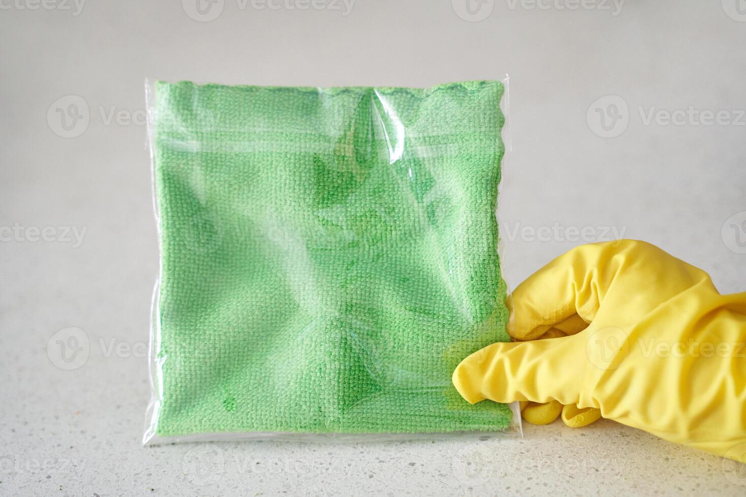Yellow gloved hand holding green cleaning cloth in plastic bag on light surface. photo