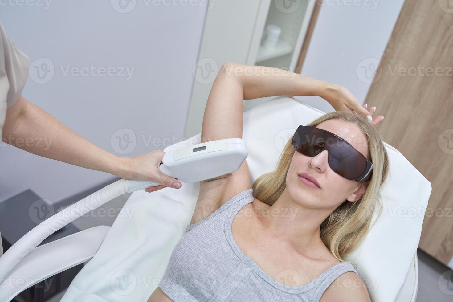 Young caucasian female undergoing laser hair removal on armpit in clinic wearing protective eyewear. photo