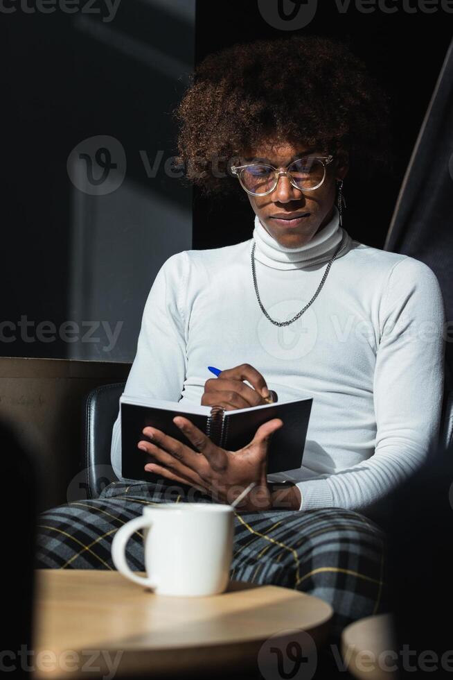 A man with glasses and a white turtleneck is sitting in a chair and writing photo