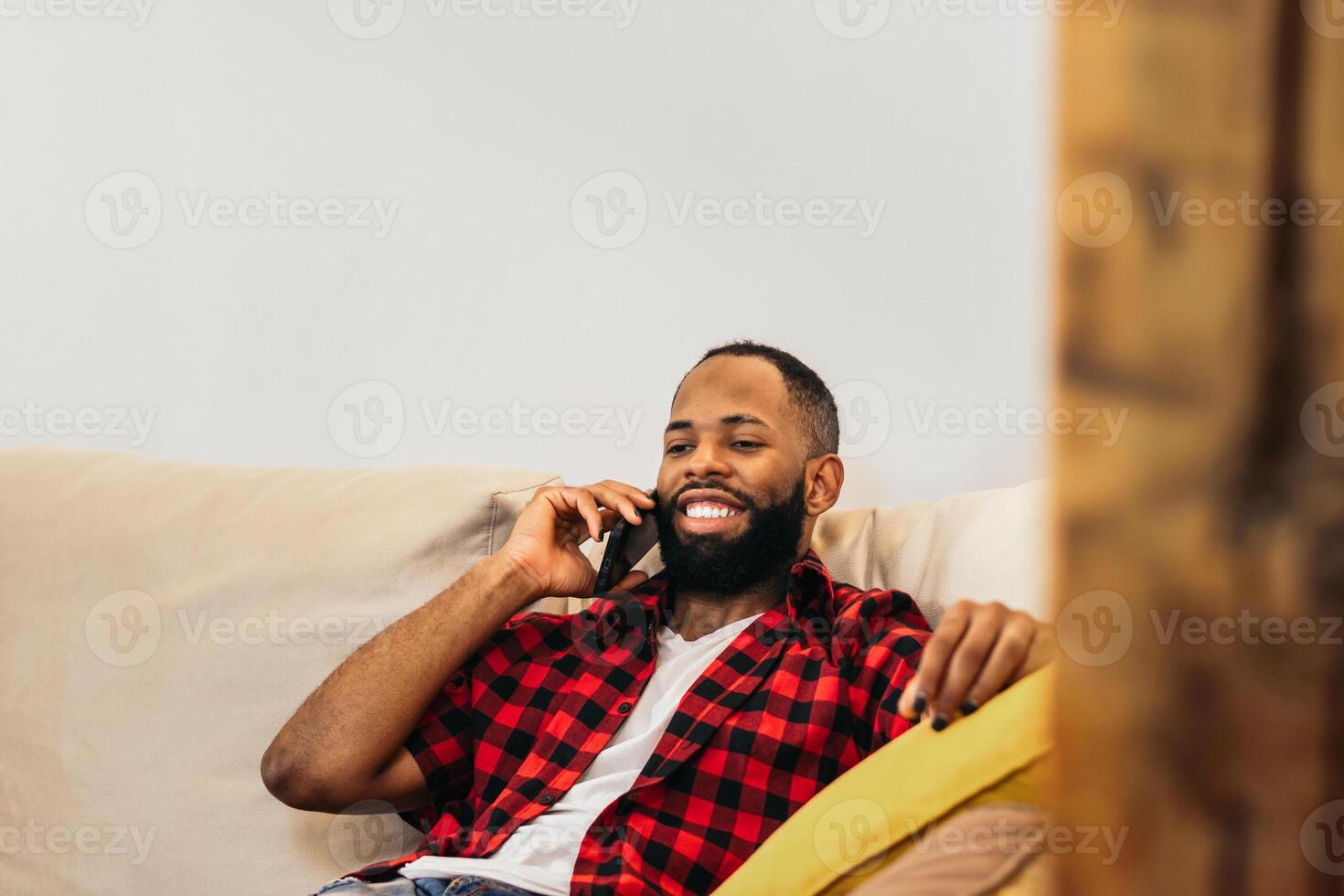 A man with a beard and checkered shirt sitting on a couch talking on his phone photo