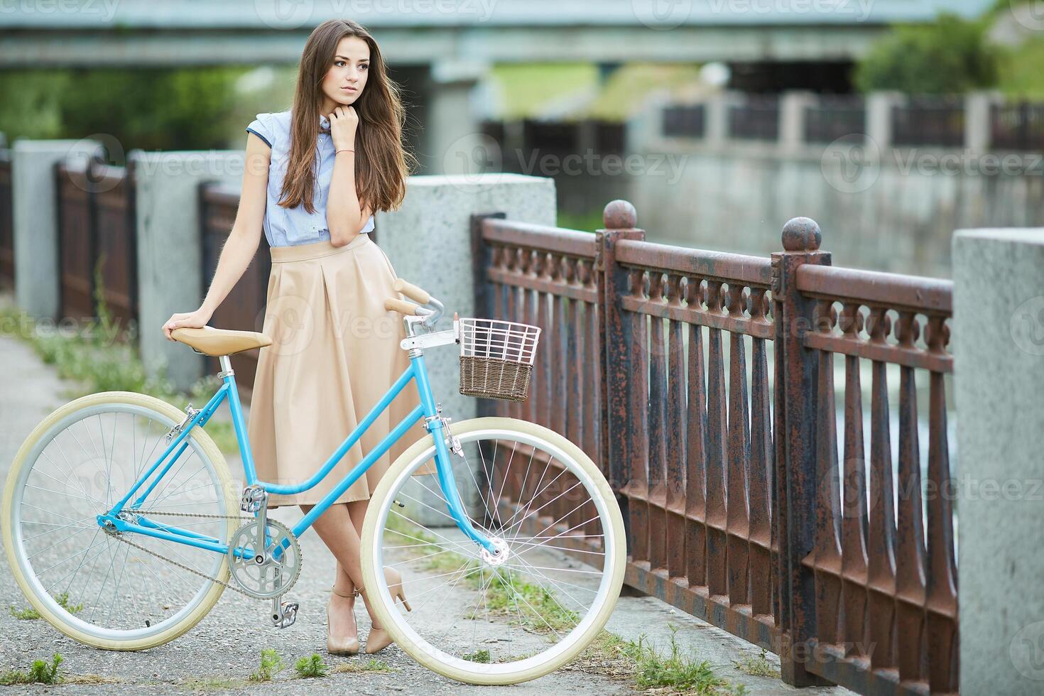 bicycle trip, Stylish young woman with long hair smiling, posing with a vintage blue bicycle on a city bridge photo