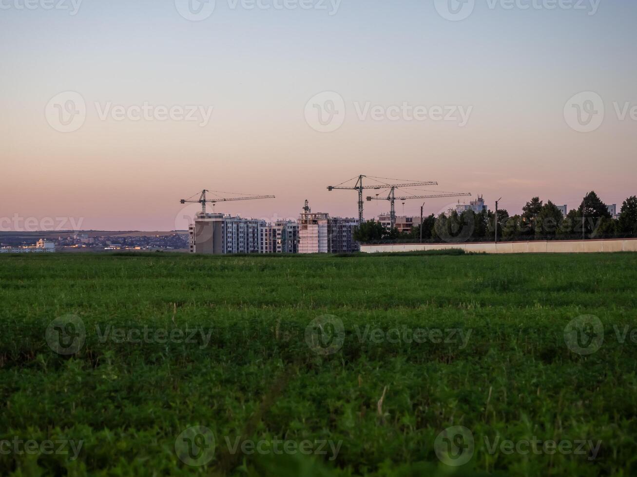 Construction project develops on the edge of a green field during twilight with cranes in the background and a peaceful atmosphere enveloping the site photo