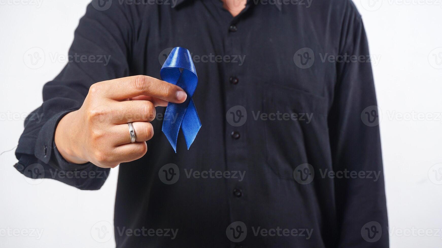 Dark blue ribbon held by man hand in front of his chest as symbol of colon cancer and colorectal cancer awareness isolated on white background photo
