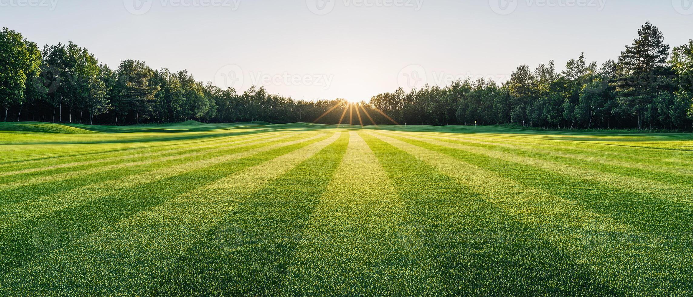 wide angle view of manicured golf course at sunrise, showcasing vibrant green grass and beautiful tree lines. sun casts rays across landscape, creating serene atmosphere photo