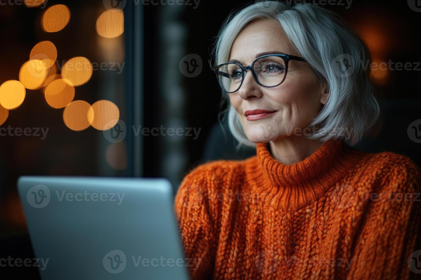 Woman in orange sweater working on laptop near window with warm bokeh lights in background photo