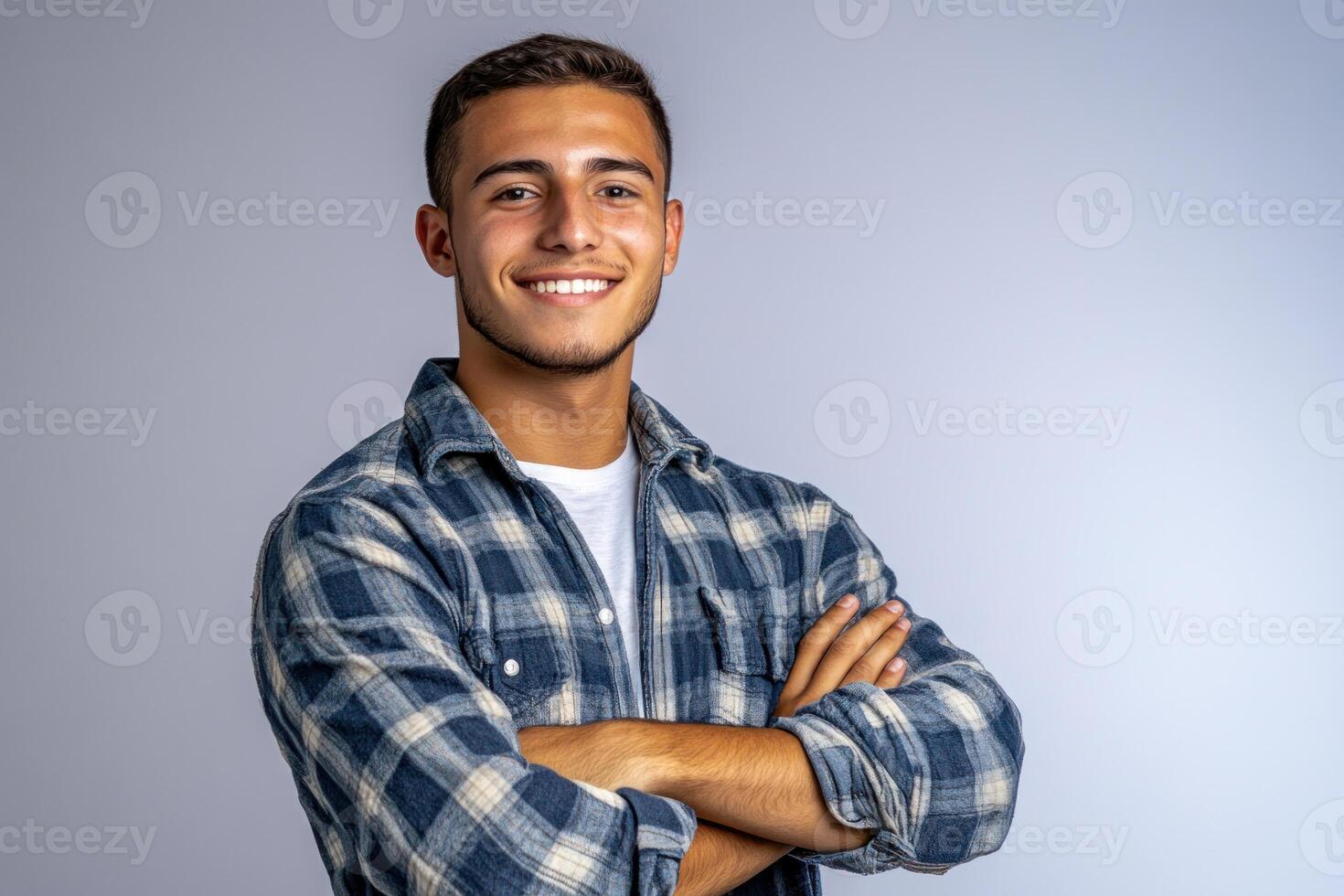 Young man smiling confidently with arms crossed wearing a checkered shirt in a studio photo