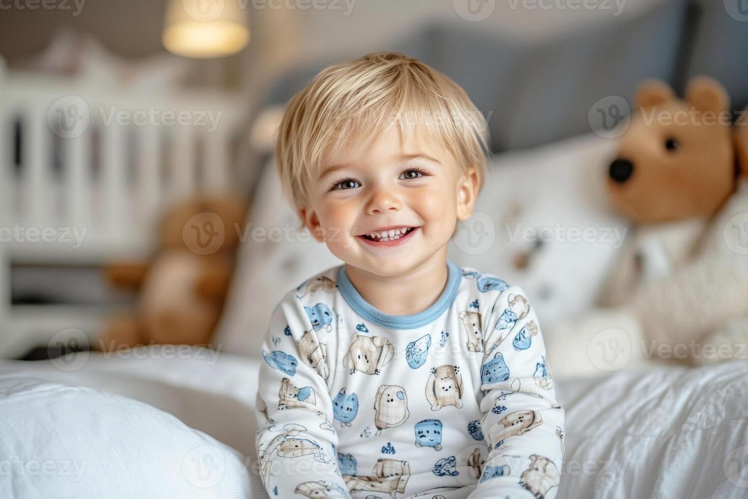 Happy blond toddler wearing pajamas with animal prints is sitting on white bed and smiling photo