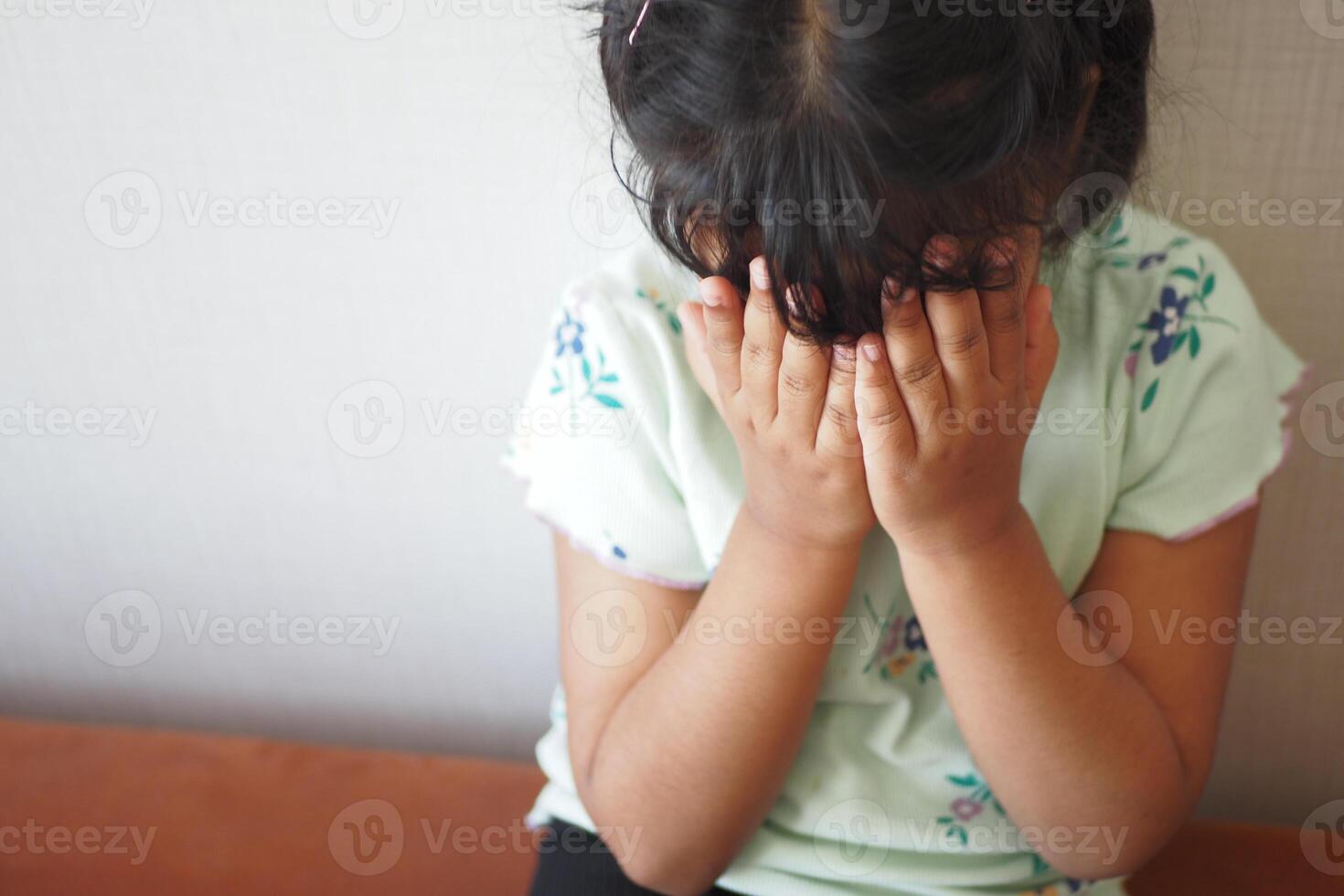 Child covering face with hands while sitting on a bench indoors photo