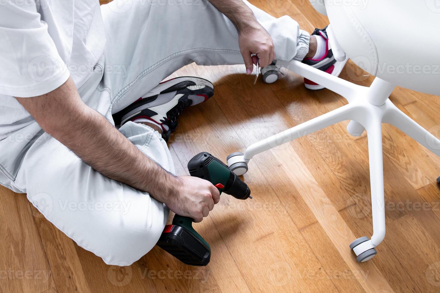 Young hispanic male assembling office chair with power drill on wooden floor photo