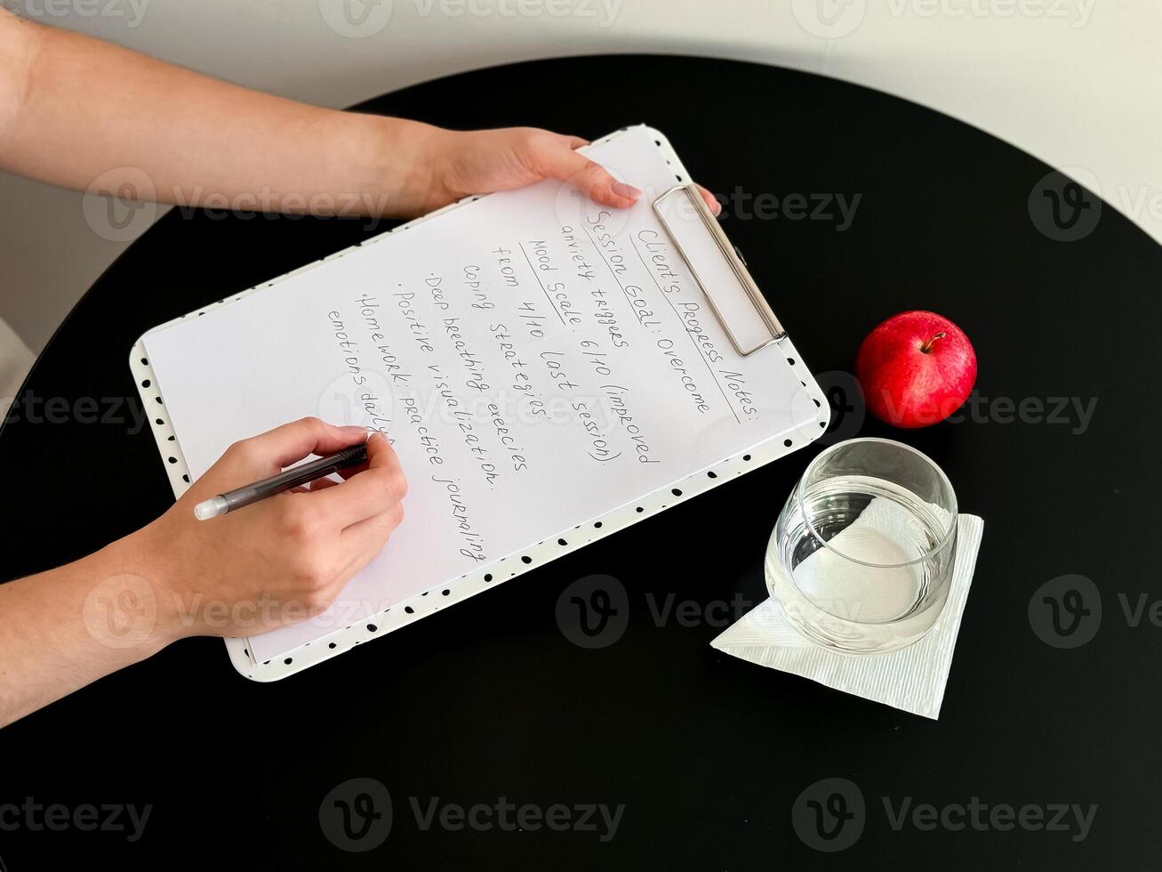 Female writing on clipboard with notes next to apple and glass on round black table photo