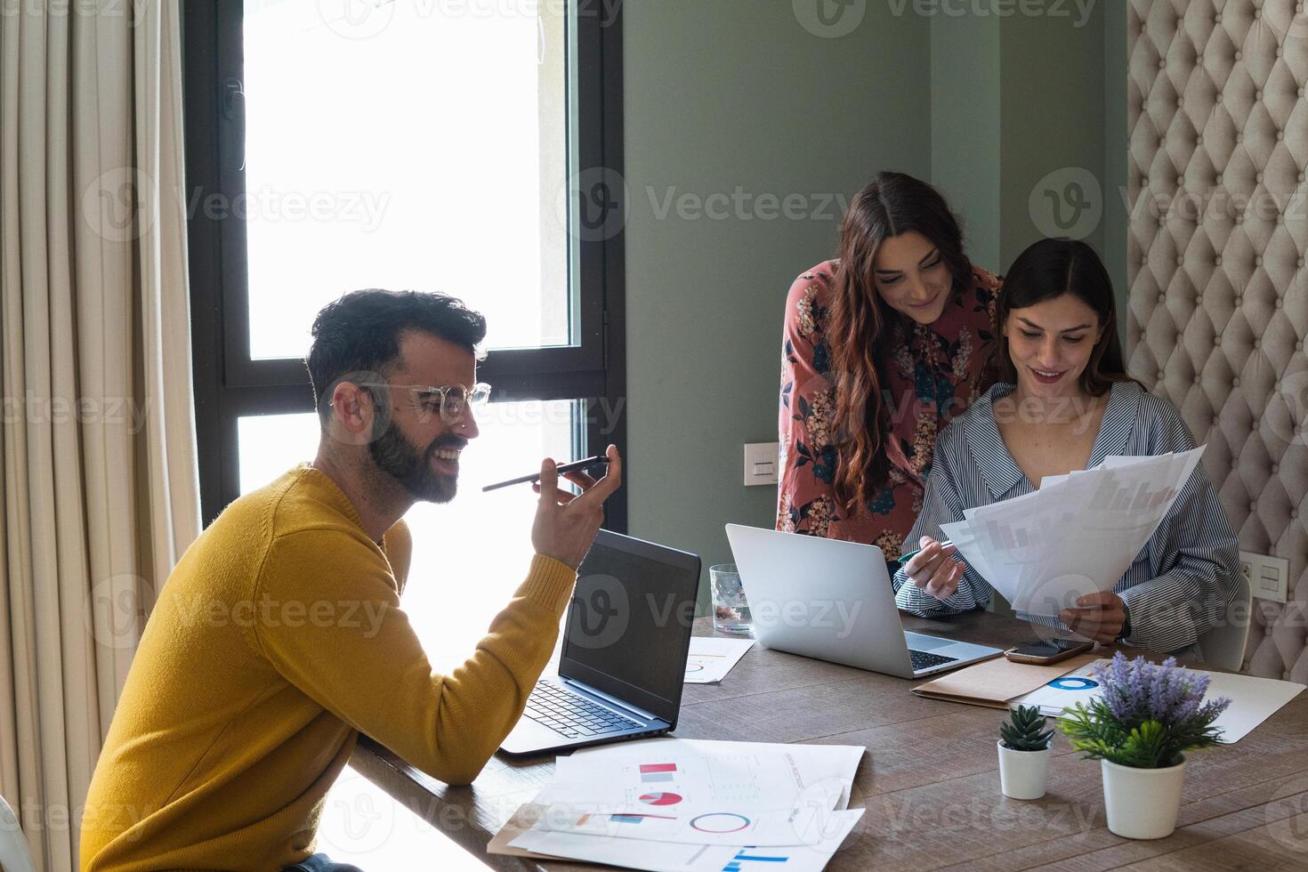 Three people sitting at a table with laptops. Coworker is having a discussion photo